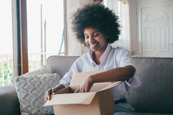 Африканская женщина сидит на диване в гостиной держать на коленях большой коробке распаковывая посылку, полученную из интернет-магазинов. Любопытная чёрная леди распаковывает картонную посылку. Раздевание женской сборной по боксу  - Фото, изображение