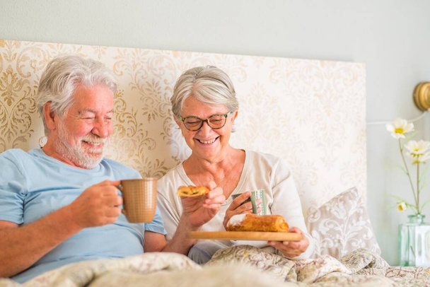 Alte ältere kaukasische Paare frühstücken morgens im Bett im Schlafzimmer zu Hause. Älteres Paar isst Croissant und trinkt Kaffee aus Tasse zum Frühstück zu Hause.   - Foto, Bild