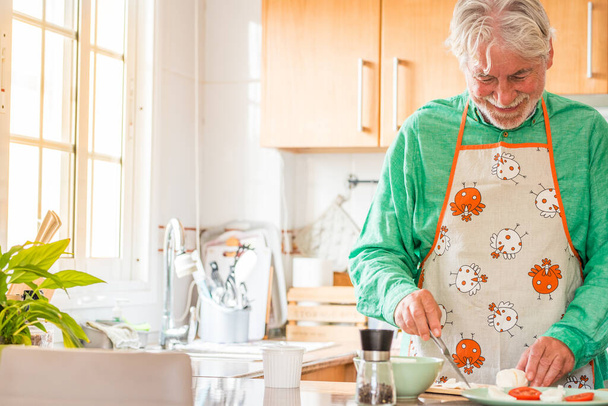 πορτρέτο ενός ώριμου και γέρου που μαγειρεύει στο σπίτι στην κουζίνα χαμογελώντας. Ο πρεσβύτερος ετοιμάζει φαγητό για μεσημεριανό μόνος του. Συνταξιούχος μάγειρας  - Φωτογραφία, εικόνα
