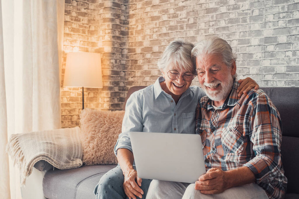 Nettes Paar alter Leute, die zusammen mit dem Laptop auf dem Sofa sitzen und einkaufen und im Netz surfen. Zwei erwachsene Menschen im Wohnzimmer genießen die Technik. Porträt von Senioren, die verliebt lachen.  - Foto, Bild