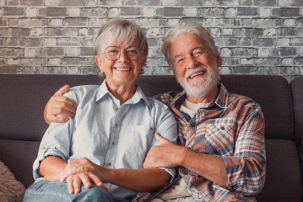 Χαριτωμένο και όμορφο ζευγάρι των ηλικιωμένων χαμογελώντας και κοιτάζοντας την κάμερα διασκεδάζοντας στο σπίτι μαζί. Πορτρέτο των ηλικιωμένων που κάθονται στον καναπέ απολαμβάνοντας και χαλαρώνοντας.  - Φωτογραφία, εικόνα