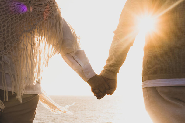 pareja de dos personas mayores felices y activas divirtiéndose y disfrutando juntos del verano en la playa caminando tomados de la mano con la puesta de sol al fondo   - Foto, imagen