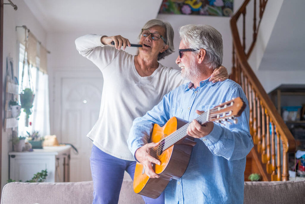 Twee gelukkige senioren of volwassen en oude mensen die samen thuis zingen en dansen. Gepensioneerde man speelt gitaar terwijl zijn vrouw zingt met een afstandsbediening van TV.  - Foto, afbeelding