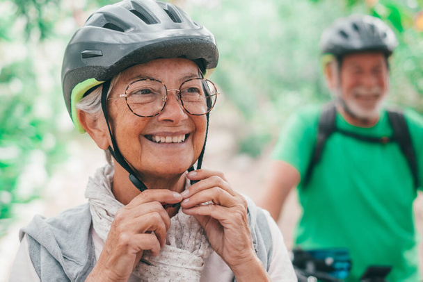 Portrait of one old woman smiling and enjoying nature outdoors riding bike with her husband laughing. Head shot of mature female with glasses feeling healthy. Senior putting on helmet to go trip with bicycles.   - Fotografie, Obrázek