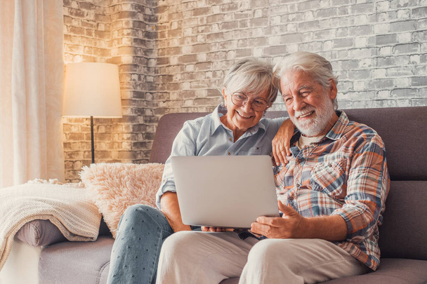 Χαριτωμένο ζευγάρι των ηλικιωμένων που κάθονται στον καναπέ χρησιμοποιώντας φορητό υπολογιστή μαζί ψώνια και σέρφινγκ στο διαδίκτυο. Δύο ώριμοι άνθρωποι στο σαλόνι απολαμβάνουν την τεχνολογία.  - Φωτογραφία, εικόνα