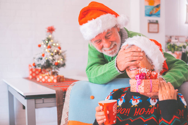 Senior couple in warm clothing and Santa hat, man covering eyes of old wife with a surprise gift for her. Loving old romantic heterosexual couple celebrating Christmas festival together  - Photo, Image