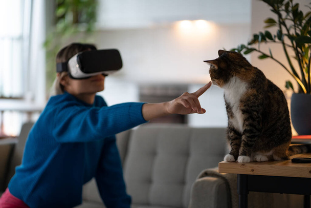 Mavi kazak giyen genç bir kadın evdeki rahat koltukta oturur ve VR gözlükleriyle oyun oynar. Dişi, evcil hayvanın kafasını okşamaya çalışır ve evde oturan kediyle oynar. - Fotoğraf, Görsel