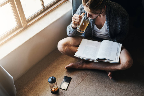 Así es como empiezo mis fines de semana. Foto de ángulo alto de un joven tomando té y leyendo un libro mientras está sentado en el suelo en casa - Foto, Imagen