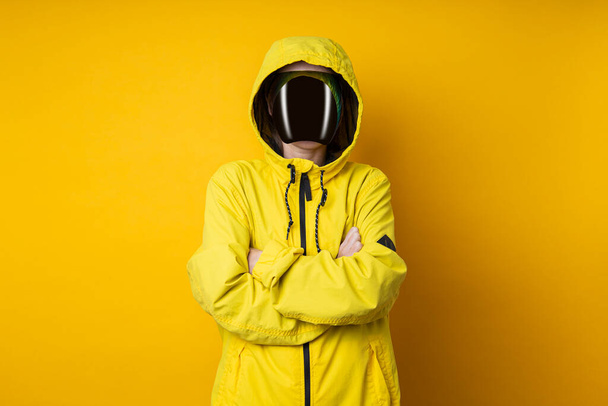 Jonge vrouw in een masker van cyberpunk bril in een geel jasje met haar armen gekruist op een gele achtergrond. - Foto, afbeelding