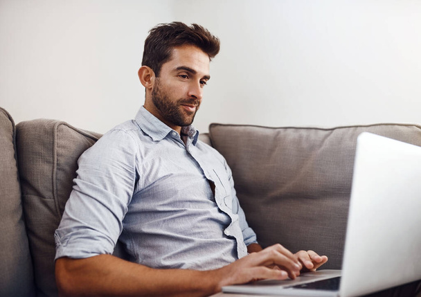 Von zu Hause aus zu arbeiten ist immer das Beste. ein fokussierter junger Mann, der an seinem Laptop arbeitet, während er tagsüber auf einer Couch zu Hause sitzt - Foto, Bild
