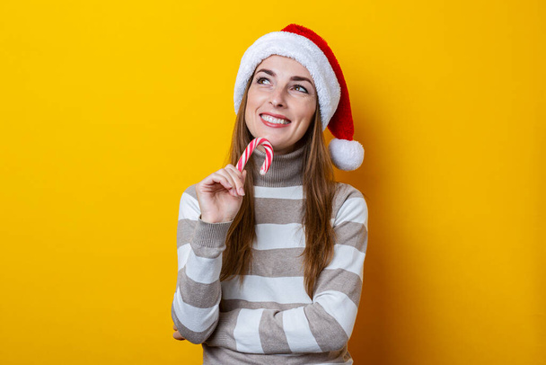 Usmívající se mladá žena v Santa Claus klobouk s vánoční cukroví na žlutém pozadí. - Fotografie, Obrázek