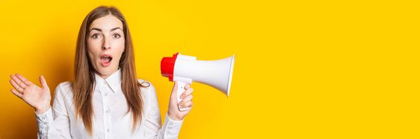 Young woman with a surprised face in a white shirt holds a megaphone in her hands on a yellow background. Hiring concept, help wanted. Banner. - Photo, Image