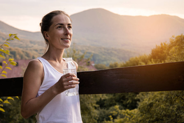 Χαμογελαστή νεαρή γυναίκα με μπουκάλι νερό απολαμβάνοντας το φυσικό τοπίο με θέα στο βουνό. - Φωτογραφία, εικόνα