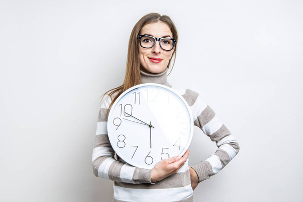 усміхнена молода жінка в окулярах тримає білий настінний годинник на світлому фоні
  - Фото, зображення