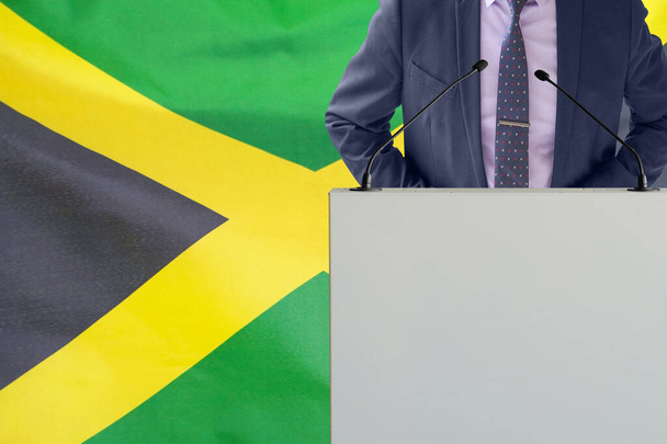 Tribune s mikrofonem a mužem v obleku na pozadí jamajské vlajky. Podnikatel a tribun na pozadí jamajské vlajky. Politik na pódiu s mikrofony na pozadí jamajské vlajky. Konference - Fotografie, Obrázek