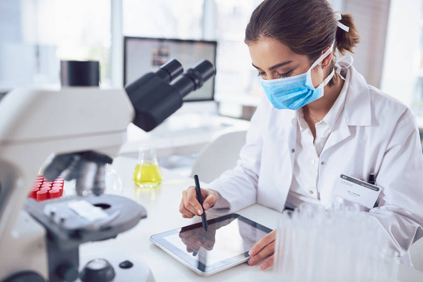 Esta tableta ha hecho que sea fácil de trabajar en el laboratorio. una joven científica enfocada usando una máscara quirúrgica y trabajando en una tableta digital dentro de un laboratorio - Foto, imagen