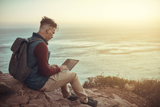 Its a great place for a bit of blogging. Full length shot of a young man using his laptop while sitting on a cliff in the mountains - Foto, Bild