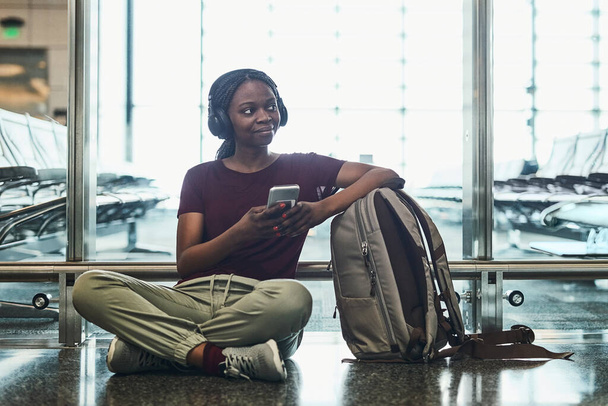 Kuuntelen musiikkia lähtöä odotellessa. nuori nainen, joka kuuntelee musiikkia puhelimellaan istuessaan lentokentällä - Valokuva, kuva