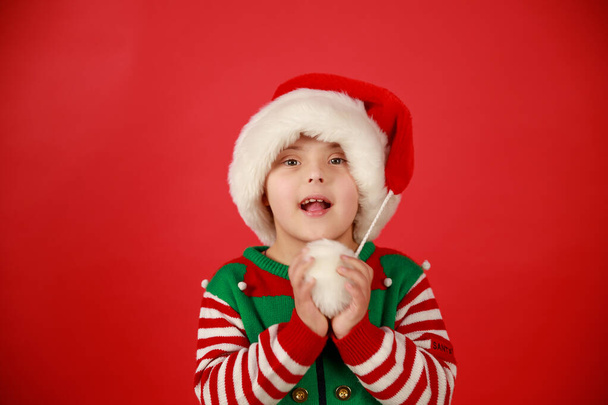 Niño feliz con síndrome de Down en un sombrero de Santa Claus y un suéter verde sobre un fondo rojo, humor navideño. Feliz Año Nuevo. - Foto, imagen