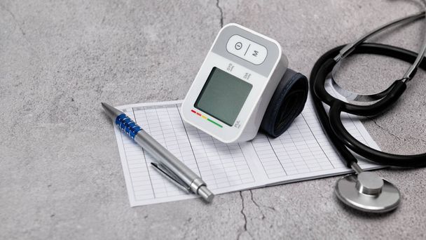 Автоматичний цифровий монітор артеріального тиску зап'ястя; діаграма моніторингу тиску; ручка; стетоскоп; на сірому фоні, банер
 - Фото, зображення