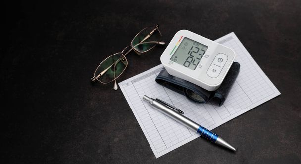 Automatický digitální monitor krevního tlaku na zápěstí, graf sledování tlaku, pero, brýle na černém pozadí - Fotografie, Obrázek