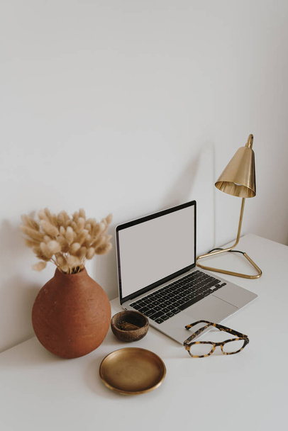 Aesthetic minimalist home office workspace desk. Laptop computer with blank copy space display screen, glasses, dried grass bouquet, lamp, golden plate. Chic lady boss business branding concept - Photo, Image