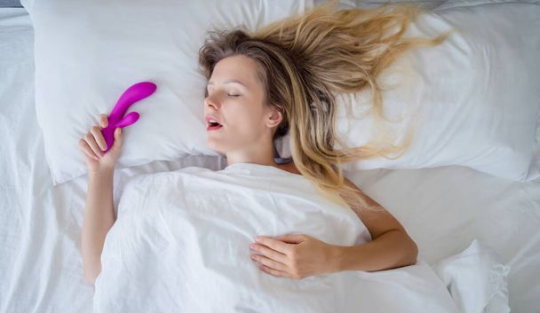 Jovem mulher sexy na cama segurando em suas mãos um brinquedo sexual para adultos. - Foto, Imagem