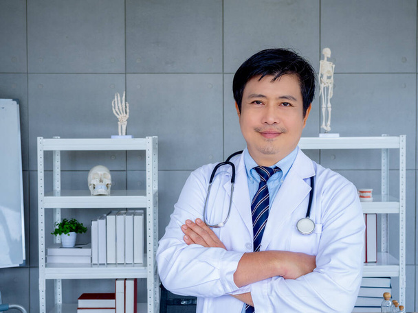 Glimlachende Aziatische man orthopedische arts portret in witte jas staan met gekruiste armen in de buurt van boekenplank en apparatuur in het medisch kantoor. Zelfverzekerde volwassen mannelijke arts of arts met stethoscoop. - Foto, afbeelding