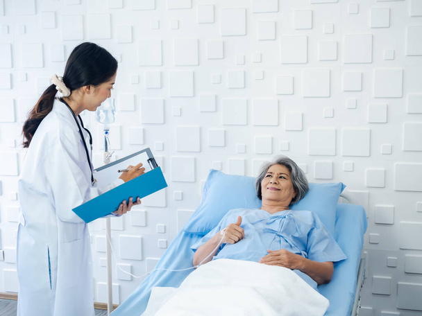 Χαμογελάστε Ασιάτισσα ηλικιωμένη γυναίκα γκρίζα μαλλιά ασθενής ξαπλωμένη στο κρεβάτι, αναρρώνει ενώ νέες γυναίκες γιατροί επισκέπτονται, εξηγώντας τα συμπτώματα και τις σημειώσεις εξετάσεων και ιατρικά έγγραφα υγειονομικής περίθαλψης στο δωμάτιο του νοσοκομείου.  - Φωτογραφία, εικόνα