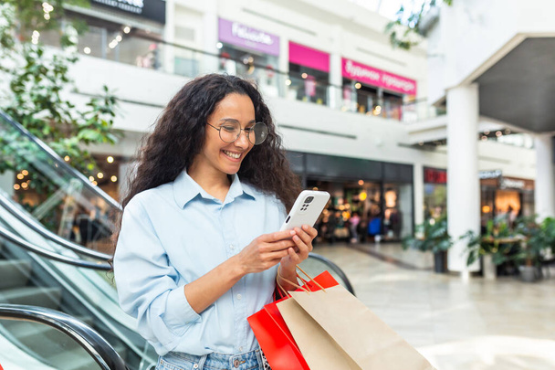 Ευτυχισμένη γυναίκα που ψωνίζει ρούχα κατά την περίοδο των εκπτώσεων και των διαφημιστικών προσφορών, Ισπανίδα γυναίκα χαμογελαστή και χαρούμενη περιήγηση σε απευθείας σύνδεση εκπτώσεις χρησιμοποιώντας smartphone, μέσα σε ένα σύγχρονο μεγάλο κατάστημα. - Φωτογραφία, εικόνα