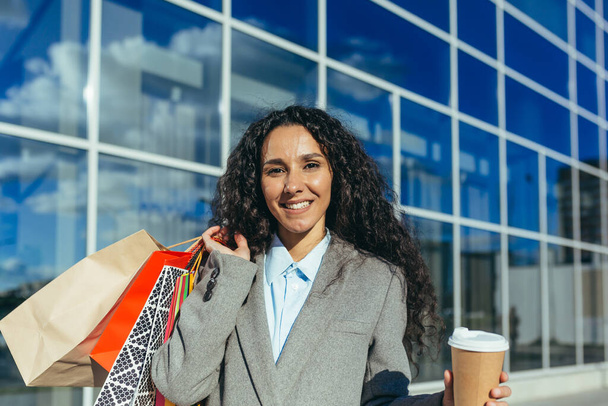 Retrato de una mujer compradora feliz, mujer hispana mirando a la cámara y sonriendo fuera de un gran supermercado, con bolsas coloridas y una taza de bebida caliente. - Foto, Imagen