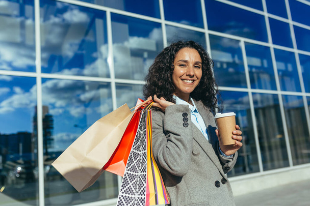 Porträt einer glücklichen Shopperin, einer hispanischen Frau, die in die Kamera blickt und vor einem großen Supermarkt lächelt, mit bunten Tüten und einer Tasse Heißgetränk. - Foto, Bild