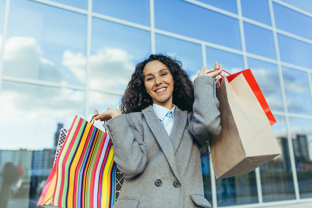 Beautiful woman with curly hair outside big store looking at camera and smiling, Hispanic woman shopper happy with discounts and sale, in warm coat portrait. - Foto, imagen