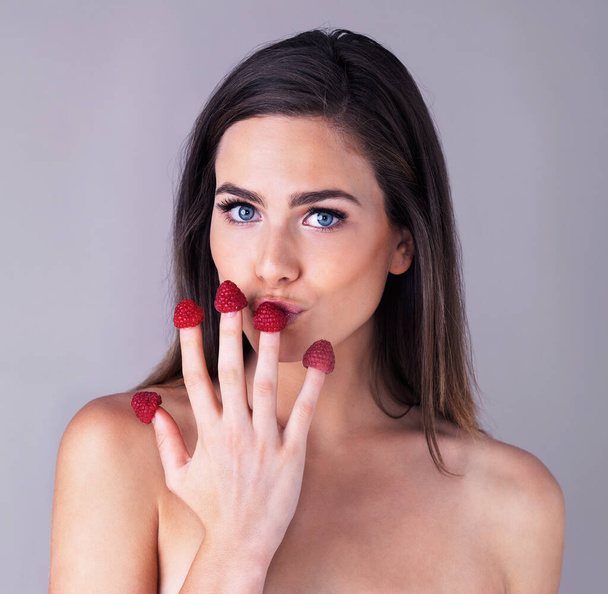 Its oh so juicy. Studio portrait of an attractive young woman eating raspberries off her fingertips against a purple background - Foto, Imagem