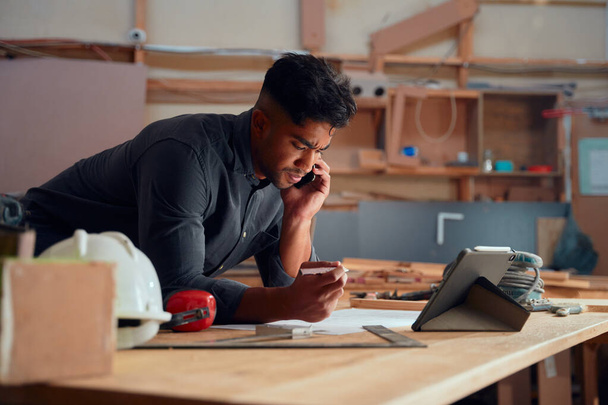 Focused multiracial young man on mobile phone leaning on elbows by digital tablet in woodworking factory - Photo, Image