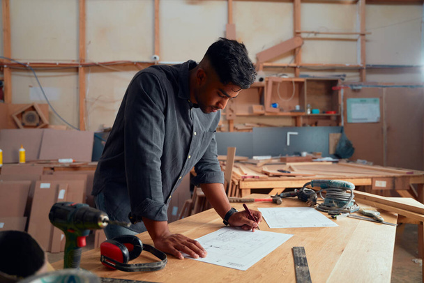 Focused multiracial young man in shirt writing with pencil on paper next to tools at woodworking factory - Foto, Bild