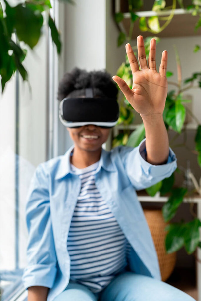 Joven mujer africana alegre en casco VR sentada en el alféizar de la ventana en casa interactuando con objetos en el mundo virtual, tocando cosas en realidad aumentada, disfrutando de una experiencia inmersiva, enfoque selectivo - Foto, imagen