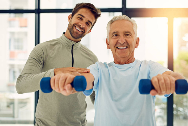 Fitness ist einfach, wenn man die richtige Hilfe hat. Porträt eines freundlichen Physiotherapeuten, der seinem älteren Patienten beim Training mit Gewichten hilft - Foto, Bild