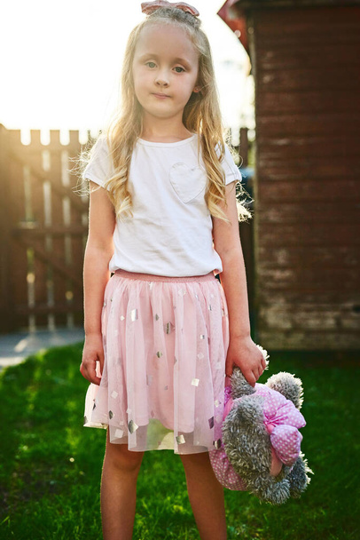 Temps de jeu dehors. Portrait de douce petite fille debout et tenant son ours en peluche tout en regardant la caméra à l'extérieur - Photo, image