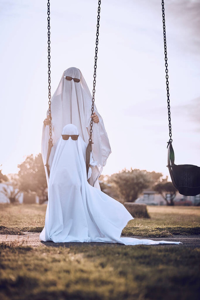 Halloween, costume and ghost couple at park, kid enjoy scary holiday with parent. Family, festival and child with mom or dad in spooky white sheet dress up with sunglasses sitting on playground swing. - Фото, зображення
