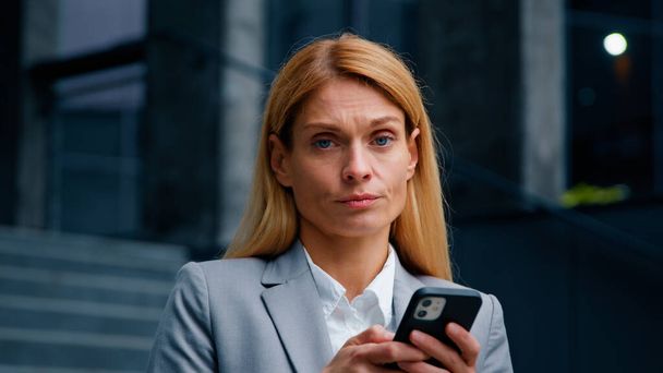 Poważny skupiony bizneswoman trzyma telefon stojąc w mieście smutny sfrustrowany kobieta odbieranie wiadomości e-mail czytanie złych wiadomości traci w zakład online zdenerwowany o utracie pieniędzy czuje nieszczęśliwe negatywne emocje - Zdjęcie, obraz