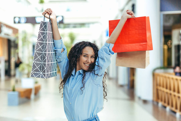Happy shopper woman looking at camera and smiling, hispanic woman with curly hair dancing and jumping with pleasure, bought gifts on sale, portrait of happy shopping woman. - Foto, afbeelding