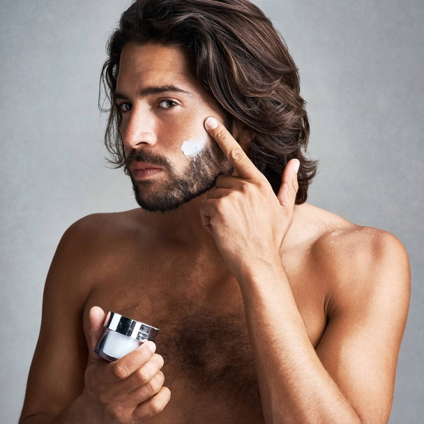 Im all about that anti ageing. Studio shot of a handsome young man applying moisturiser to his face against a grey background - Φωτογραφία, εικόνα