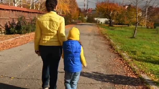 Вид на брюнетку в жёлтой куртке и синих джинсах, любящую мать, держащую дочь за руки, идущую по улице вместе в прекрасный тёплый солнечный осенний день. Выходные вместе - Кадры, видео