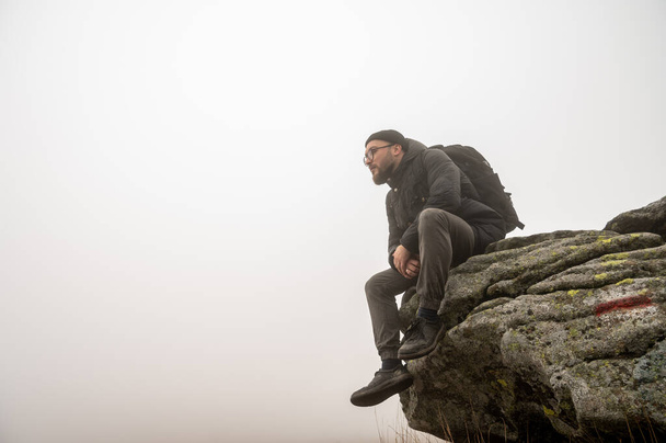 Egy szemüveges, szakállas, utazó fiatalember ül a hegy szélén, köd, köd, köd, pihenés a hegyekben. Szabad hely az íráshoz - Fotó, kép