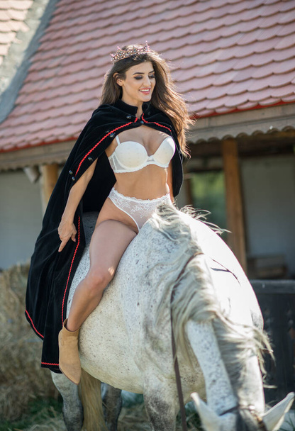 sensual brunette princess with sexy white bikinis riding a horse . Portrait of a girl  with cape and crown and her horse. Girl interacting and having fun with a horse in the  story fairytale style - 写真・画像