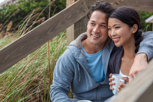 Young Asian Chinese man and woman, boy & girl, dating couple sitting on wooden steps overlooking a beach drinking mugs of tea or coffee - Photo, Image