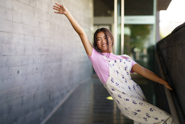 Gelukkig jong aziatisch vrouw in stijlvolle outfit verhogen arm en glimlachen terwijl grijpen helling reling en kantelen terug buiten modern gebouw - Foto, afbeelding