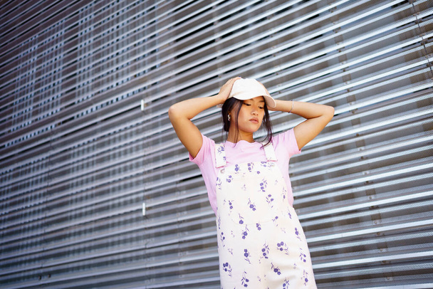 Genç Asyalı kadın şık giyinmiş, şapkalı, kafasına dokunmuş ve modern binanın metal duvarının yanında dikilirken aşağıya bakıyor. - Fotoğraf, Görsel