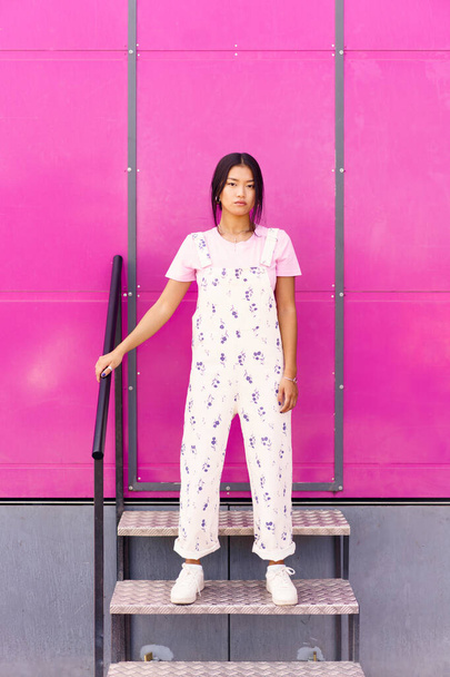 Cuerpo completo joven mujer asiática en ropa elegante tocando barandilla y mirando a la cámara mientras está de pie en los pasos contra la pared rosa - Foto, Imagen